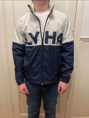 Helly Hansen jacka  - Snygg vindjacka i blått och vitt från helly hansen med stor logga över bröst och rygg. Jackan har dragkedja framtill, hög krage och elastiska muddar vid ärmsluten. Perfekt för blåsiga dagar och enkel att matcha till jeans.