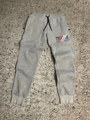 Gråa Trapstar mjukisbyxor - Säljer ett par gråa mjukisbyxor från Trapstar med broderad text i blått, rött och vitt på ena benet. Byxorna har svarta dragsnören i midjan, muddar vid bensluten och sidofickor. Perfekta för en chill och avslappnad stil.