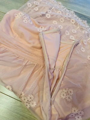 Ljusrosa axelbandslös spetsklänning - Söt ljusrosa axelbandslös klänning med blommig spets och dragkedja i sidan. Klänningen har fina broderade detaljer längs hela tyget. 