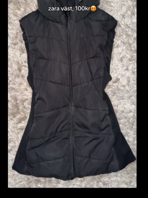 Svart Zaraväst - Snygg svart dunväst från Zara med huva och dragkedja framtill. Västen har en quiltad design och är perfekt för lager-på-lager-stil. Tillverkad i ett mjukt och vadderat material som håller dig varm under kyliga dagar!