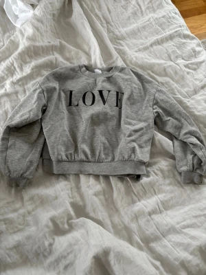 Grå sweatshirt med LOVE-tryck🩶 - Säljer en grå sweatshirt med svart LOVE-tryck på bröstet. Tröjan har rund halsringning, långa ärmar och ribbade muddar vid ärmslut och nederkant. Perfekt för en chill och avslappnad stil. Materialet känns mjukt och bekvämt mot huden.🌸
