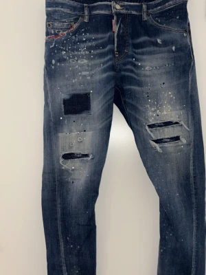 DSQUARED2 Jeans - Säljer ett par blå jeans med coola slitningar, patchdetalj och färgstänk. Jeansen har en relaxed passform och klassisk femficksdesign från DSQUARED2. Perfekta för dig som gillar en edgy och avslappnad stil. 