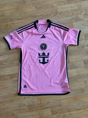Rosa Inter Miami matchtröja Adidas - Snygg rosa matchtröja från Inter Miami CF med svarta detaljer och klubbmärke på bröstet. Tröjan har Adidas-logga, svarta ränder på axlarna och tryck på ärmen. Tillverkad i lätt och ventilerande funktionsmaterial, perfekt för fotboll eller träning.