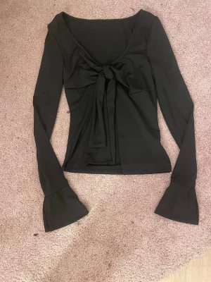 Svart långärmad topp med knyt - Trendig svart långärmad topp med snygg knytning framtill och utsvängda ärmslut. Toppen har en djupare ringning och är figurnära i modellen, perfekt för att lyfta din outfit. Materialet känns mjukt och stretchigt.