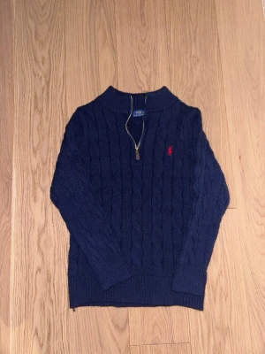 Mörkblå kabelstickad tröja Polo Ralph Lauren - Mörkblå kabelstickad tröja från Polo Ralph Lauren med halv dragkedja och röd broderad logga på bröstet. Tröjan har ribbade muddar och hög krage, perfekt för kyliga dagar. Klassisk och tidlös design som passar till det mesta.