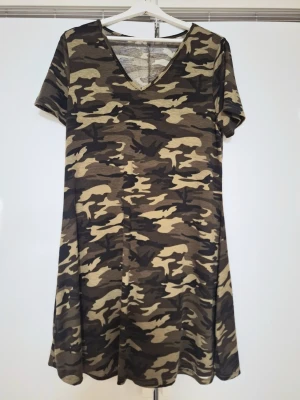 Camouflagemönstrad klänning  - Cool kort klänning med camouflagemönster i gröna, beige och bruna toner. Klänningen har v-ringning och korta ärmar, perfekt för en avslappnad och trendig look. Tillverkad i mjukt och stretchigt material för extra komfort. Passar även XL. Aldrig använd men lapparna är bortklippta. 