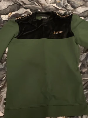 Grön och svart tröja från Vichy - Snygg grön tröja från Vichy med svart sammetsliknande panel upptill och guldfärgad logga på bröstet. Dekorativa detaljer på axlarna. Perfekt för dig som vill sticka ut med en unik stil.