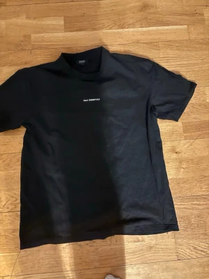 Svart t-shirt från SMOG - Säljer en svart t-shirt från  SMOG med trycket 'Daily Essentials' på bröstet och en stor grafisk ruta i svart, grått, vitt och rött på ryggen. Klassisk passform och rund hals. Perfekt för dig som gillar stilrena men ändå unika plagg.