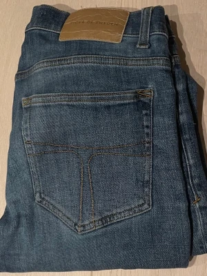 Blå jeans från Tiger of Sweden - Snygga blå jeans från Tiger of Sweden med klassisk femficksmodell och raka ben. Jeansen har en diskret tvättad look och kontrastsömmar. Tillverkade i mjukt denimtyg som ger en skön passform. Perfekta för en avslappnad och stilren outfit.
