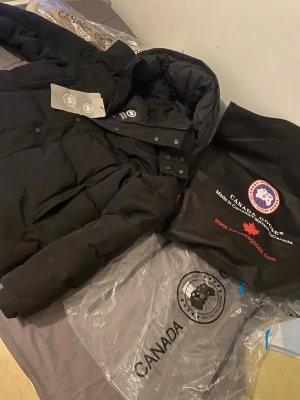 Svart dunjacka från Canada Goose - Säljer en svart dunjacka från Canada Goose med avtagbar pälskant till huvan. Jackan har klassisk quiltad design, hög krage och tryckknappar. Perfekt för kalla vinterdagar och riktigt varm. Märkeslogga på ärmen och flera fickor.