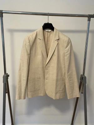 Beige linnekavaj med klassisk krage - Stilren beige kavaj i linne med klassisk krage och två framfickor. Enkelknäppt modell med rak passform, perfekt för en avslappnad men ändå uppklädd look.