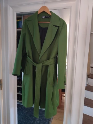 Grön kappa med skärp från Zara - Stilren grön kappa från Zara med klassisk krage och knytskärp i midjan. Kappan har två stora fickor framtill och är helfodrad med svart innerfoder. Perfekt för dig som vill sticka ut med färg i höst och vinter.
