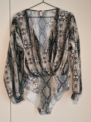 Ormmönstrad body med lång ärm - Säljer en snygg body med ormmönster i svart, grått och vitt. Djup omlott-ringning framtill och långa puffiga ärmar med resår vid handlederna. Tillverkad i mjukt, stretchigt material. Perfekt för dig som vill sticka ut med ett trendigt plagg.