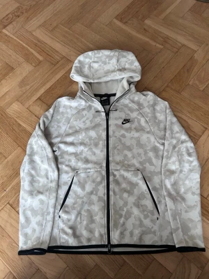 Nike hoodie med camomönster och dragkedja - Snygg hoodie från Nike i ljusbeige och vitt camomönster. Tröjan har hel dragkedja framtill, två fickor med svarta dragkedjor och svart Nike-logga på bröstet. Huva och svarta detaljer vid ärmslut och nederkant. Perfekt för en sportig och avslappnad stil.