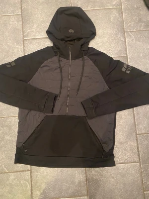 Svart hoodie från Sail Racing - Säljer en svart hoodie från Sail Racing med quiltad front, dragkedja halvvägs och stor huva med dragsko. Logga på både ärm och huva. Hoodien har en stor magficka och är tillverkad i en mix av bomull och syntetmaterial för extra komfort.