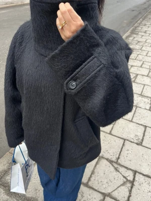 JACKA I ULL- OCH ALPACKABLANDNING MED STÅKRAGE - Säljer du en svart ull och alpackablandad kappa med hög krage och knäppning framtill. Jackan har en bred passform, stora fickor och dekorativa knappar vid ärmsluten. Kappan är slutsåld hos Arket! Storlek 36!