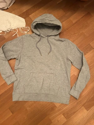 Grå basic hoodie med ficka - Säljer en stilren grå hoodie med huva och dragsnören. Hoodien har en stor magficka framtill och är gjord i mjukt bomullsmaterial. Perfekt för en avslappnad och trendig look.