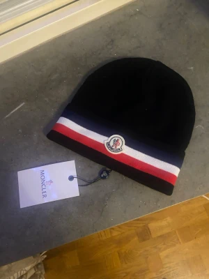 Svart Moncler mössa med ränder - Snygg svart mössa från Moncler med klassiska ränder i rött, vitt och blått runt kanten. Mössan har Moncler-loggan framtill och är tillverkad i mjukt, stretchigt material som sitter skönt på huvudet. Perfekt accessoar för att lyfta din outfit.