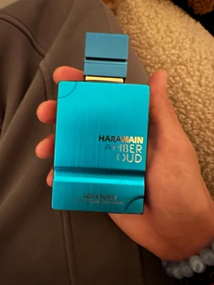 Haramain Amber Oud Aqua Dubai parfym - Håller 10+ timmar Snygg parfymflaska i metalliskt blått från Haramain, modellen Amber Oud Aqua Dubai. Flaskan har en modern, fyrkantig form med gulddetaljer och matchande blått lock. Perfekt för dig som gillar exklusiva och fräscha dofter. Använd fåtal gånger 