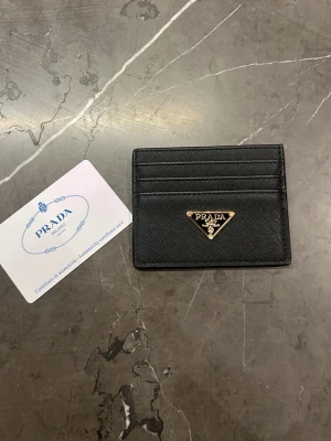 Svart kortfodral från Prada - Stilrent svart kortfodral från Prada i saffianoläder med guldfärgad metallogo framtill. Fodralet har flera kortfack och en elegant, minimalistisk design som passar perfekt i fickan. Klassisk accessoar för dig som gillar lyxiga detaljer.