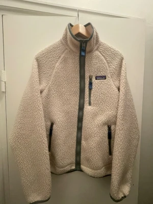 Fleecejacka från Patagonia - Mysig beige teddyjacka från Patagonia med hög krage och dragkedja framtill. Jackan har tre fickor med dragkedja och detaljer i grått. Tillverkad i fluffigt teddy-material som håller dig varm och bekväm. Perfekt för kalla dagar och snygg till jeans eller leggings.