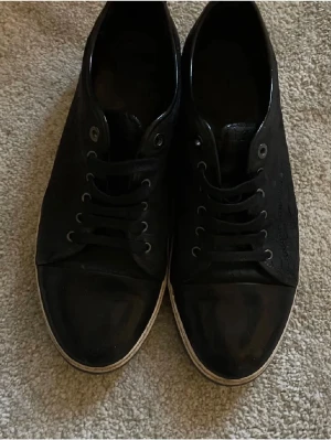Svarta Lanvin sneakers i läder med croc mönster. - Snygga svarta sneakers från Lanvin med ovandel i läder och klassisk låg siluett. Skorna har svarta skosnören, metallöglor och en vit sula med diskret logga undertill. Perfekta för dig som gillar stilrena och exklusiva sneakers.