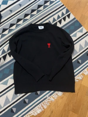 Svart stickad tröja från Ami - Svart stickad tröja från Ami med rund halsringning och ribbade muddar. På bröstet finns Ami:s ikoniska röda hjärta med bokstaven A. Tröjan har en klassisk passform och är perfekt för dig som gillar stilrena detaljer.