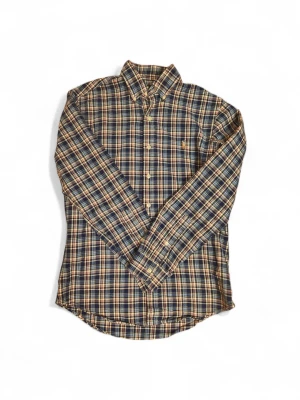 Rutig skjorta från Polo Ralph Lauren - Klassisk rutig skjorta från Polo Ralph Lauren i blått, beige och gult. Skjortan har lång ärm, knappar framtill och en liten broderad logga på bröstet. Tillverkad i mjuk bomull och har en tidlös passform som funkar till många stilar.