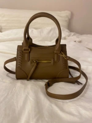 Brun minihandväska med axelrem - Snygg brun minihandväska i slätt konstläder med guldfärgade detaljer. Väskan har både handtag och justerbar axelrem samt en liten ficka med dragkedja framtill. Dekorativa sömmar längs kanterna ger en cool touch. Perfekt för dig som gillar stilrena accessoarer.