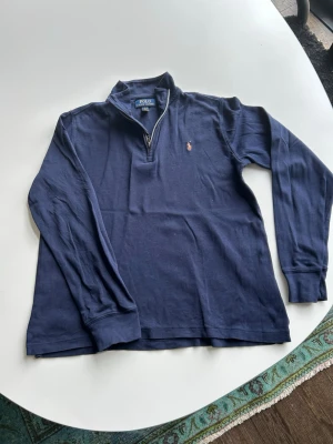 Marinblå halvzip från Ralph Lauren L 14-16 år - Snygg marinblå långärmad tröja från Polo Ralph Lauren (ungdom) med halv dragkedja och klassisk krage. Tröjan har det ikoniska broderade logotypen på bröstet och är tillverkad i mjuk bomull. Perfekt för en avslappnad men stilren look. Liten i storleken. 