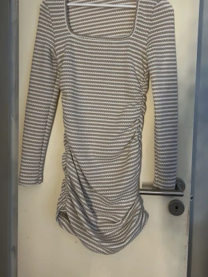Randig beige och vit klänning - Snygg kortklänning med beige och vita ränder, trekvartsärm och rynkade detaljer på sidan. Klänningen har en fyrkantig ringning och är gjord i ett mjukt stretchigt material. Perfekt för dig som gillar en stilren men ändå unik look. Endast testad