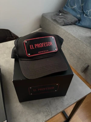 Svart keps El Profesor John Hatter - Snygg svart keps från John Hatter & Co med mesh på sidorna och bak. Framtill sitter en metallskylt med texten 'EL PROFESOR' i rött och vitt. Justerbar snapback och böjd skärm för en cool och avslappnad look.