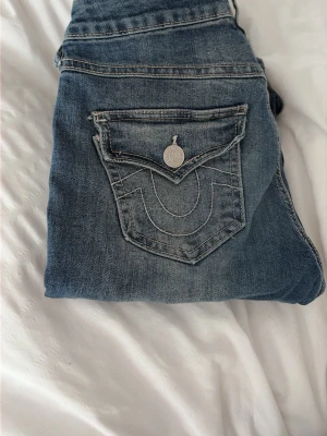 Blå jeans från True Religion - Säljer ett par klassiska efterfrågade blå jeans från True Religion med deras ikoniska bakfickor och kontrastsömmar. Jeansen är skräddar syd för att passa 160-163 cm, tyvärr är de för små för mig och saknar en knapp.  PRIS KAN DISKUTERAS 