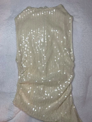 Beige paljett-topp med asymmetrisk kant - OANVÄND En beige topp täckt med glittrande paljetter och en  asymmetrisk nederkant. Ärmlös modell med hög halsringning och dragkedja i ryggen. Perfekt för dig som vill sticka ut med en trendig och festlig look.