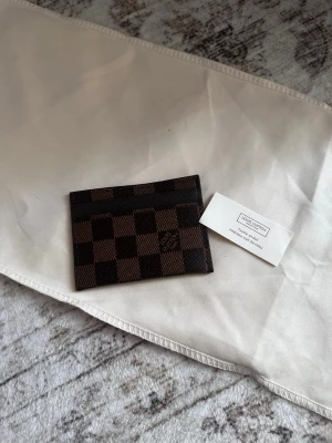 Louis Vuitton brun kortfodral - Snyggt kortfodral från Louis Vuitton i klassiskt brunt damier-mönster. Tillverkat i tåligt canvasmaterial med plats för flera kort. Perfekt accessoar för dig som gillar stilrena och lyxiga detaljer.