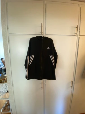 Svart Adidas hoodie med dragkedja - Svart hoodie från Adidas med vit logga på bröstet och klassiska vita ränder längs ärmarna. Tröjan har dragkedja framtill och en skön huva. Perfekt för en sportig och avslappnad stil. Materialet känns mjukt och bekvämt.