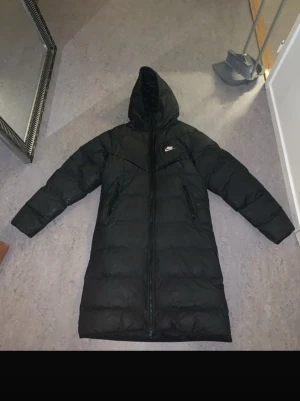 Svart lång dunjacka från Nike - Snygg svart lång dunjacka från Nike med huva och dragkedja hela vägen. Jackan har två sidofickor med dragkedja och klassisk Nike-logga på bröstet. Perfekt för kalla vinterdagar och riktigt varm tack vare dunfyllningen.  Storlek xs men passar s 