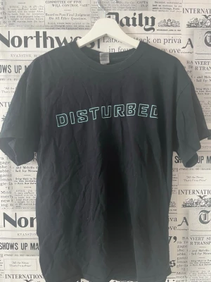 Svart Disturbed t-shirt från Gildan - Säljer en svart t-shirt från Gildan med trycket 'DISTURBED' i blått på bröstet. Klassisk rund hals och korta ärmar. Tillverkad i kraftig bomull, perfekt för dig som gillar bandmerch och vill ha en enkel men snygg look. Se måttet i bilderna 