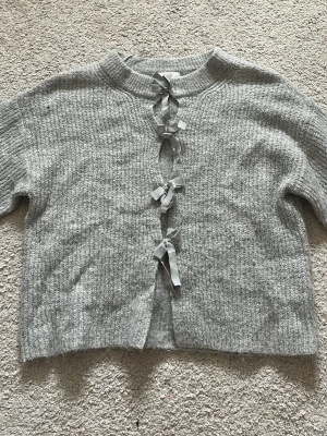 Grå kofta med rosetter Gina Tricot - Supersöt grå stickad kofta från Gina Tricot med fyra dekorativa rosetter framtill. Koftan har ribbade muddar och en croppad passform. Perfekt för dig som gillar gulliga detaljer och vill ha något mysigt att slänga på sig.