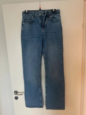 Blå raka jeans med hög midja - Säljer ett par klassiska blå jeans med rak passform och hög midja. Jeansen har fem fickor, bälteshällor och stängs med knapp och dragkedja.