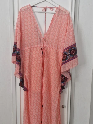 Lång korallfärgad mönstrad klänning med knyt - Långklänning i ljusrosa med vitt mönster och dekorativa bårder i mörkare färger på ärmar och nederkant. Klänningen har v-ringning, snörning framtill och i nacken. Vida, korta ärmar. Perfekt för sommarens varma dagar. Onesize men skulle säg 36-42. Ny endast provad