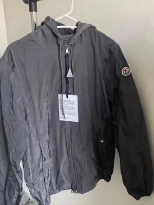 Grå vindjacka från Moncler - Snygg grå vindjacka från Moncler med huva och dragkedja framtill. Jackan har Moncler-logga på ärmen och en cool detalj med röd, vit och blå rand på huvan. Tillverkad i lätt och tåligt material, perfekt för blåsiga dagar.