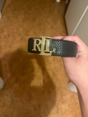 Svart skärp från Ralph Lauren - Snyggt svart skärp från Ralph Lauren med guldfärgat RL-spänne. Bältet är gjort i präglat skinn och har en klassisk, stilren look som passar till det mesta. Perfekt accessoar för att lyfta din outfit. Ralph Lauren märket är lite slitet men inte nånting man tänker på om man inte kollar noga.