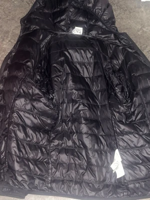 Svart tunn  dunjacka från EA7 Armani  - Snygg svart dunjacka från EA7 med quiltad design och huva. Jackan har dragkedja framtill och två sidofickor. Perfekt för kyligare dagar och ger en sportig look.