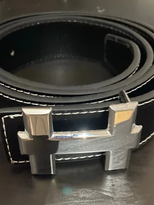 Svart skärp med silverfärgat H-spänne - Snyggt svart bälte med ett stort, silverfärgat H-format spänne och vita kontrastsömmar längs kanterna. Perfekt accessoar för att lyfta din outfit med en stilren och modern touch.