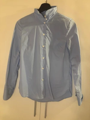 Ljusblå skjorta från H&M - Snygg ljusblå skjorta från H&M med klassisk krage och vita knappar framtill. Skjortan har lång ärm och är tillverkad i bomull, perfekt för en clean och stilren look. Använt en gång och inte mer då den inte kommer till användning💕