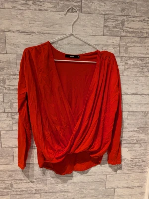 Röd omlottopp från Bik Bok - Snygg röd omlottopp från Bik Bok med djup v-ringning och långa ärmar. Toppen har en lös passform och är tillverkad i ett mjukt tyg som ger ett avslappnat intryck. Perfekt att styla med jeans eller kjol.