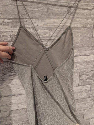 Glittrig silver kortklänning - Säljer en glittrig silverfärgad kortklänning med tunna axelband och v-ringning.. Klänningen har en figurnära passform och smala korsade band i ryggen för en snygg detalj. Perfekt för dig som vill sticka ut med en trendig look.