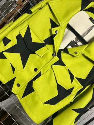 Gul jeansjacka med svarta stjärnor - Cool croppad jeansjacka i neon-grön med stora svarta stjärnor över hela jackan. Klassisk krage, två bröstfickor med lock och silverfärgade knappar. Perfekt statement-plagg för dig som vill sticka ut och endast uppsydd i ett fåtal ex