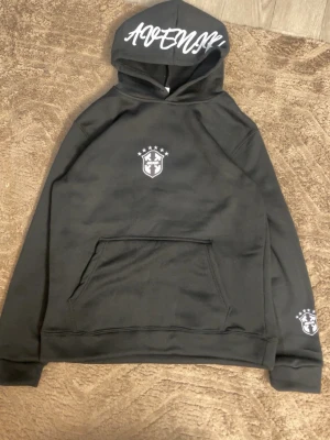 Saint Brazilian hoodie - Svart hoodie från Saint med vit text på huvan och ett stilrent emblem på bröstet samt ärmen. Klassisk känguruficka framtill och mjukt material som känns skönt mot huden. Perfekt för en avslappnad streetwear-look.